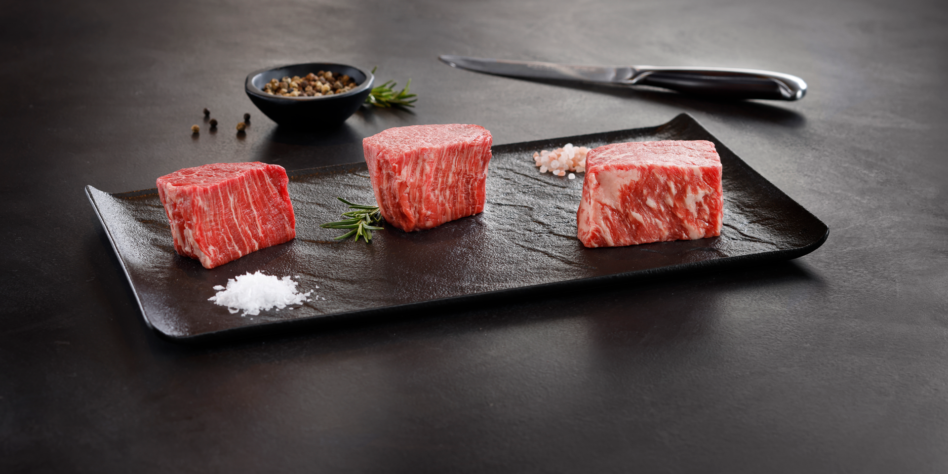 A sleek black rectangular plate displays three cuts of richly marbled Wagyu beef, showcasing the distinct marbling and high quality of the meat. The plate is garnished with sprigs of fresh rosemary, coarse salt, and a small dish of peppercorns, while a sharp knife rests in the background on a dark, elegant surface. The presentation emphasizes the luxurious and premium nature of the Wagyu beef.